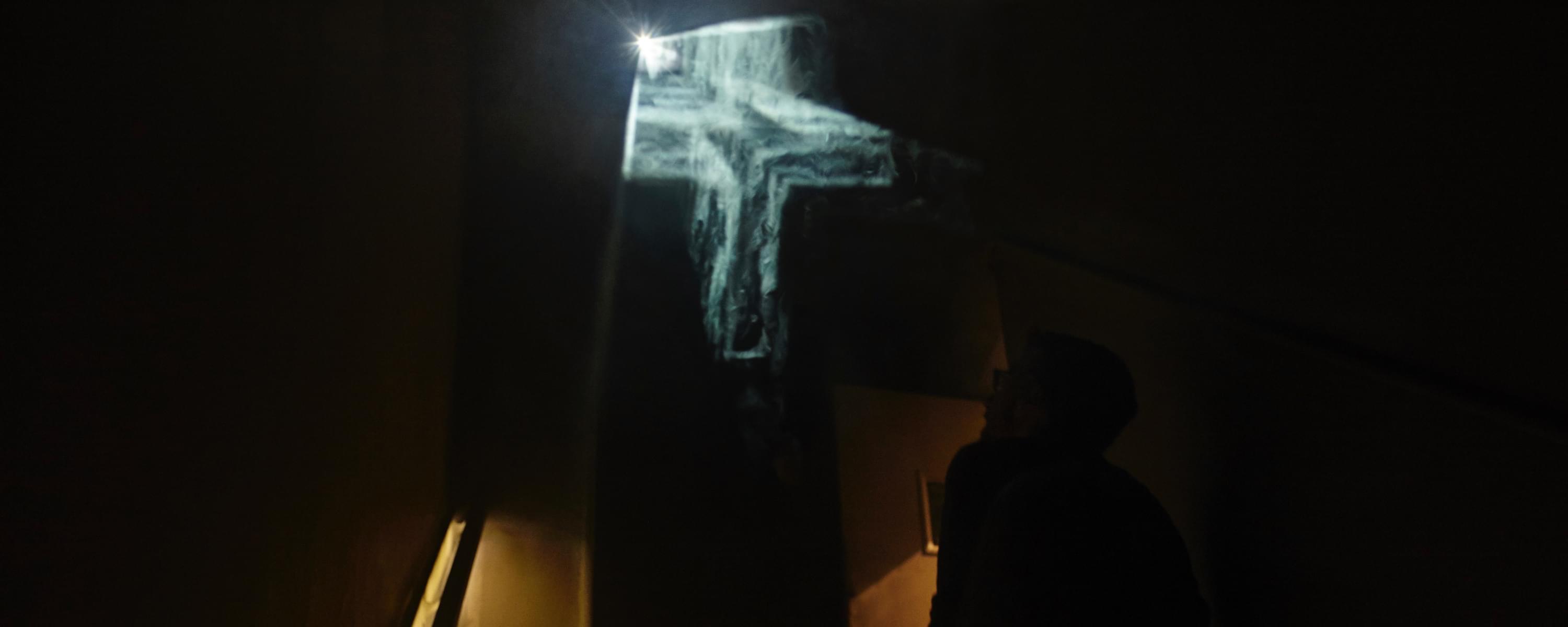 Photograph; it is extremely dark, on the right is an inkling of an illuminated handrail indicating a staircase. In the middle is a floating, cloudy, illuminated crucifix in three dimensions; towards the bottom right, if one squints, one can make out a viewer in the darkness looking up at the ghostly crucifix
