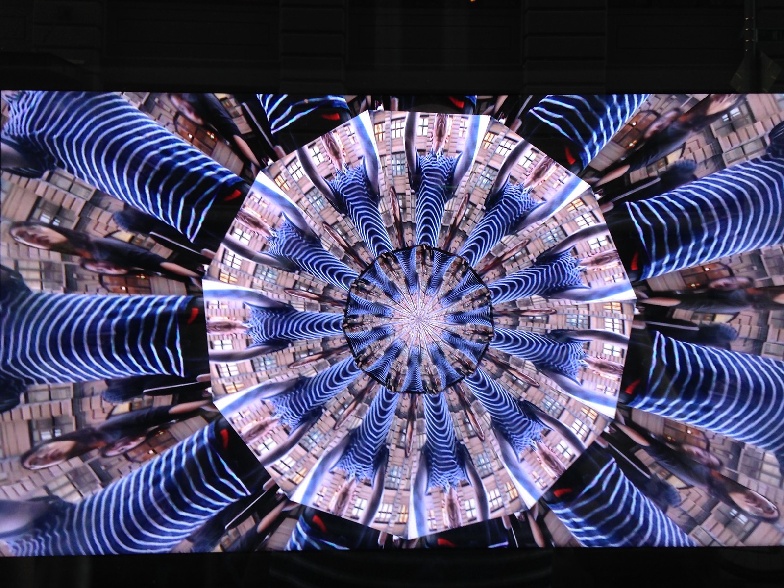 Photograph of a screen showing a kaleidoscope of infinite-mirror-esque concentric discs, each one subdivided into multiple tranches that show the same image, albeit rotated by a consistent amount across the disc. In this case, the image that gets repeated across all discs is a person’s black and white striped t-shirt, lending the overall image a very zebra-stripe-like quality.