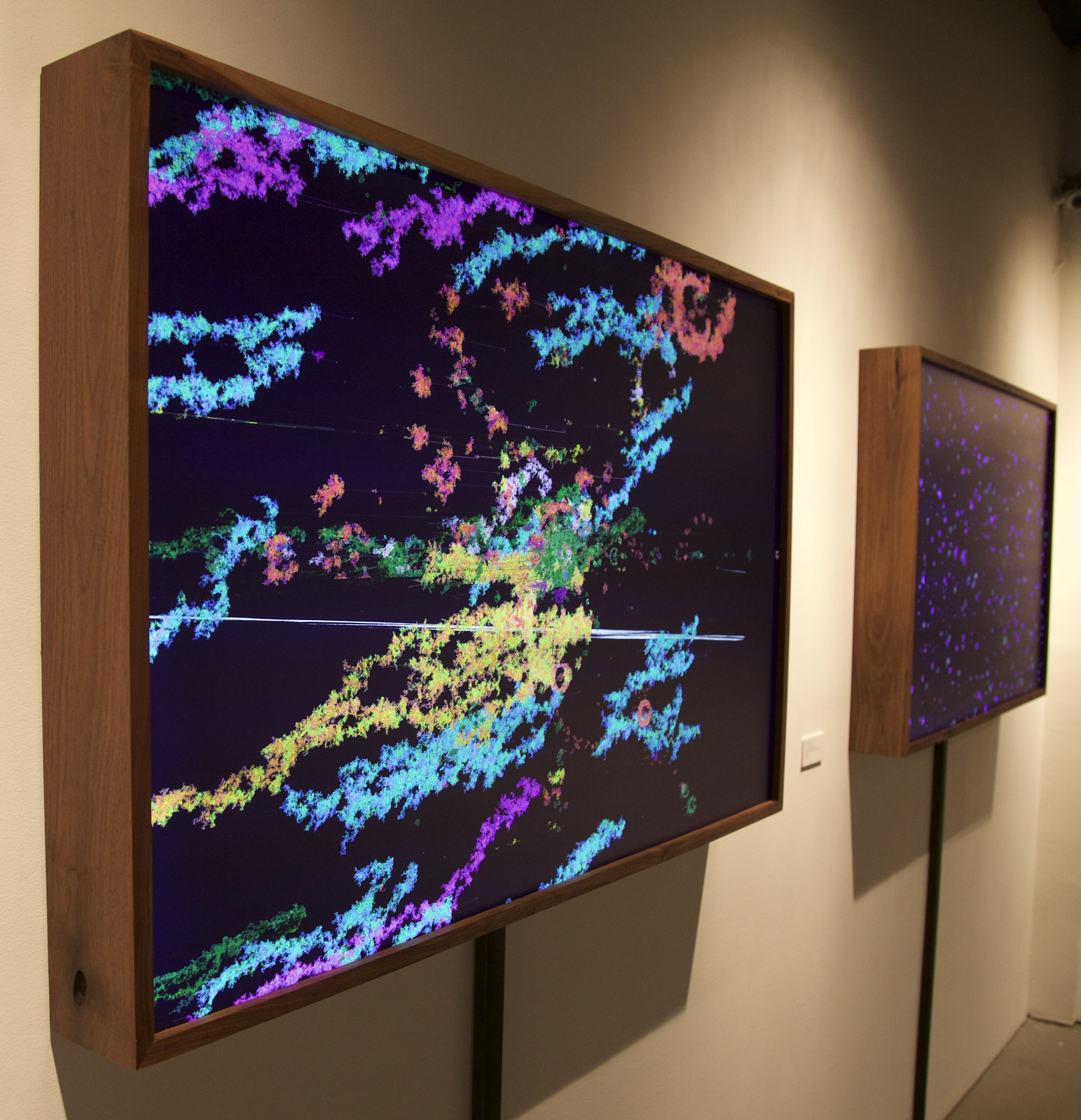 Photograph of the inside of a gallery that shows two landscape-oriented works of art in wooden frames hanging on a wall. The photo is shot from a roughly 45-degree angle.