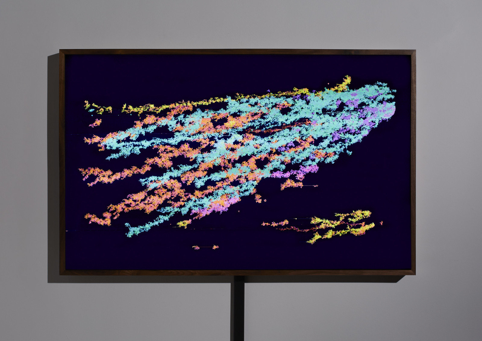 Photograph of a wooden-framed, illuminated artwork hanging on a gray wall showing concentrated masses of teal and turquoise pixel clusters, seemingly expanding from left to right, against a deep blue background.