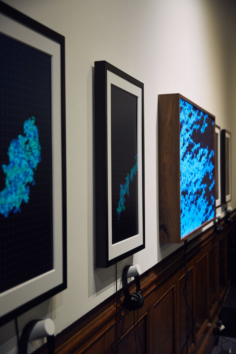 Close-up photograph of three teal blue artworks hung on a white wall, taken at a 45-degree angle such that the black metal frames are prominently shown.