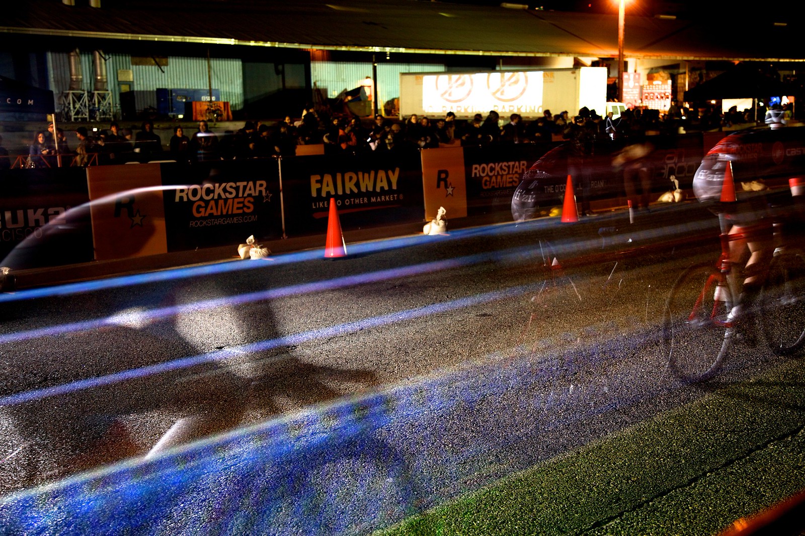 Nighttime, long-exposure photograph showing the “light lines” of two racing cyclists with a large crowd behind them. Onto the street on which they race is projected a linearly oriented rainbow road of colored lanes.
