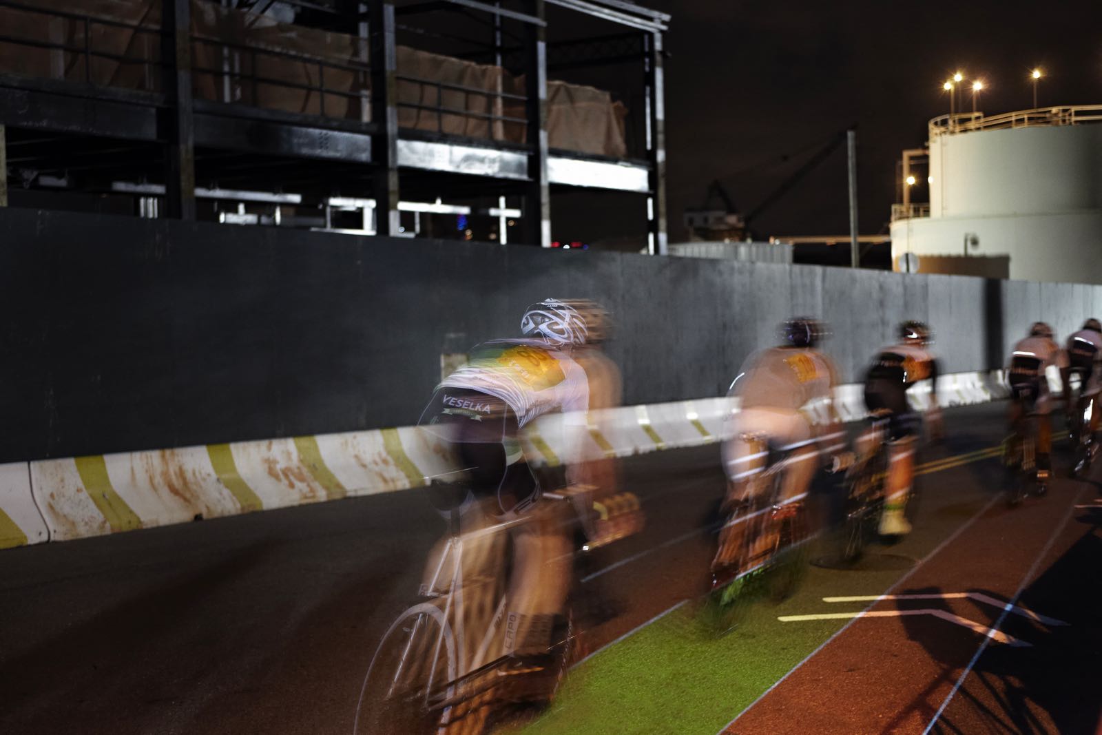 Nighttime, long-exposure photograph showing the “light lines” of five racing cyclists with a large crowd behind them. Onto the street on which they race is projected a linearly oriented rainbow road of colored lanes
