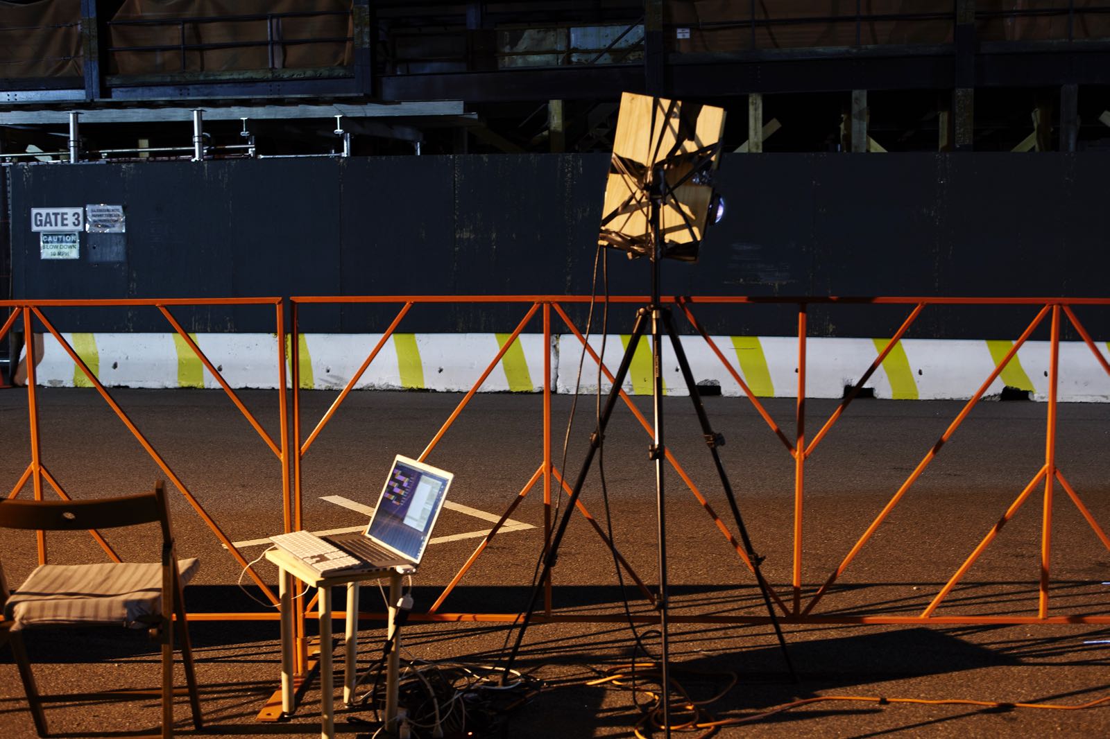 Nighttime photograph showing an orange racing barrier/fence that spans the full length of the frame. In front is a chair with an open laptop on a stand connected to a projector on a high tripod. In the background is an empty race course. There aren’t any people in the photograph.