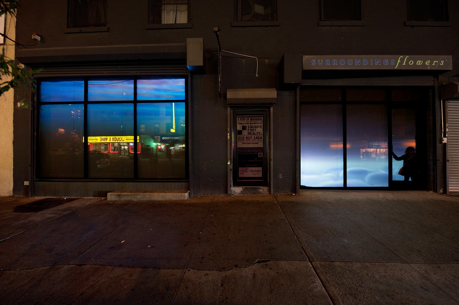 Nighttime photograph showing a wide, urban sidewalk in the foreground and a wide storefront that occupies the full width of the frame in the background. The storefront consists of two large windows on either side separated by a door in the middle. Projected into the leftmost window is an image showing water from beneath the surface and projected in the rightmost window is a purple-hue image of a sky with fluffy clouds effectively resting at sidewalk level. There is a black silhouette of a toddler wearing a dress in front of the rightmost window.