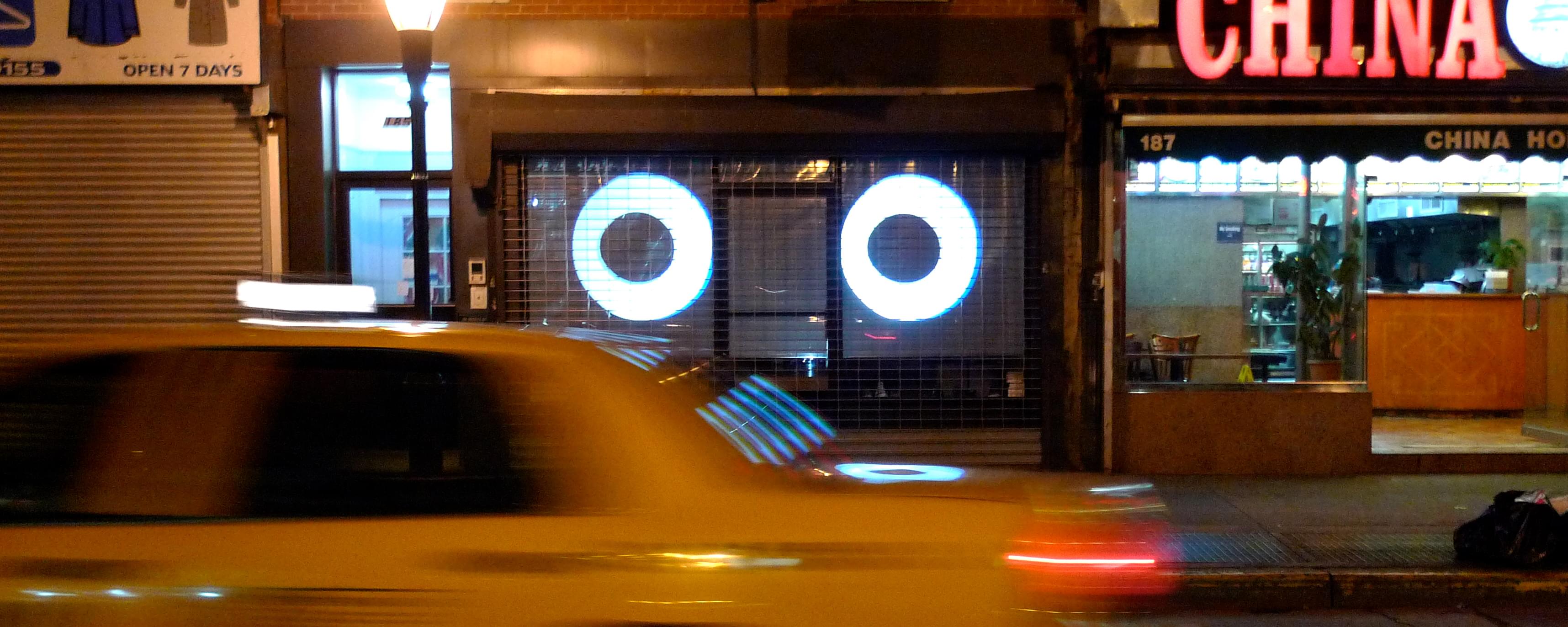 Nighttime photograph of two large, cartoonish eyes projected into a storefront with a metal security gate in the background. In the foreground, a NYC yellow cab whizzes by.